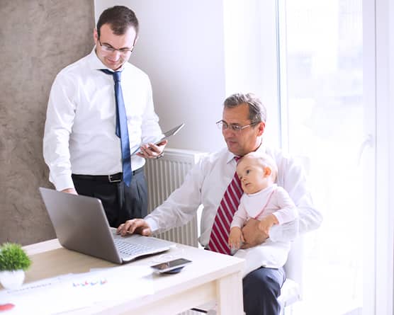 Two people working together at desk, one holding baby while reviewing laptop