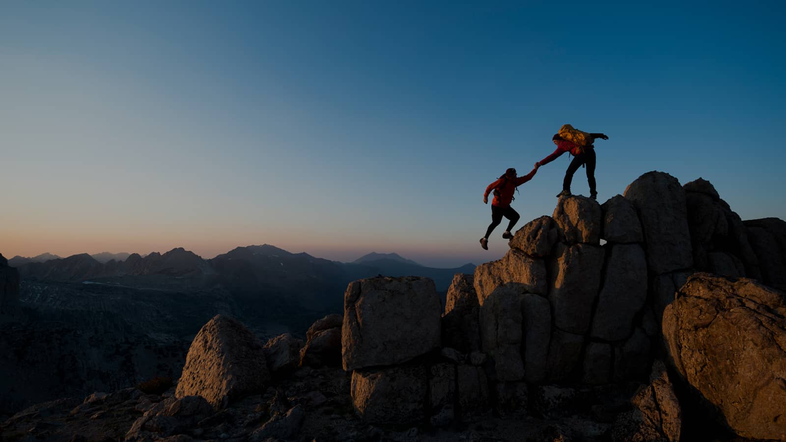 Two hikers helping each other climb rocky mountain peak at sunset Two hikers helping each other climb rocky mountain peak at sunset