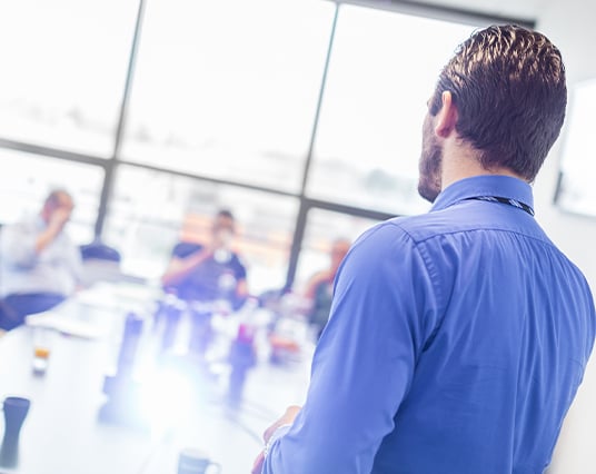 Man in blue shirt viewed from behind observing team meeting in bright modern office Man in blue shirt viewed from behind observing team meeting in bright modern office